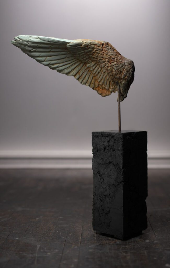 A bird sculpture perched on a slender rod attached to a black block.