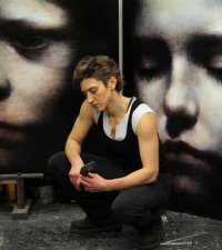 A man sitting pensively in front of large, somber portraits of faces.