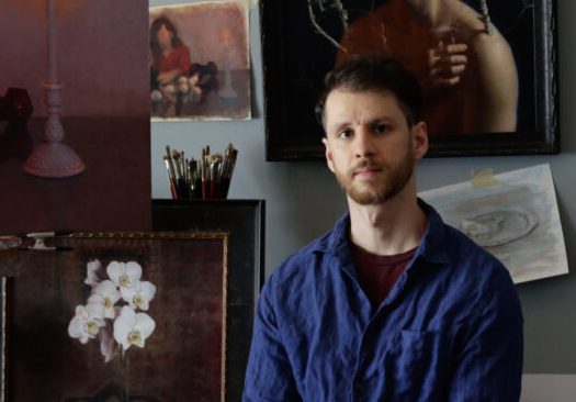 A man posing in an art studio with paintings and brushes.