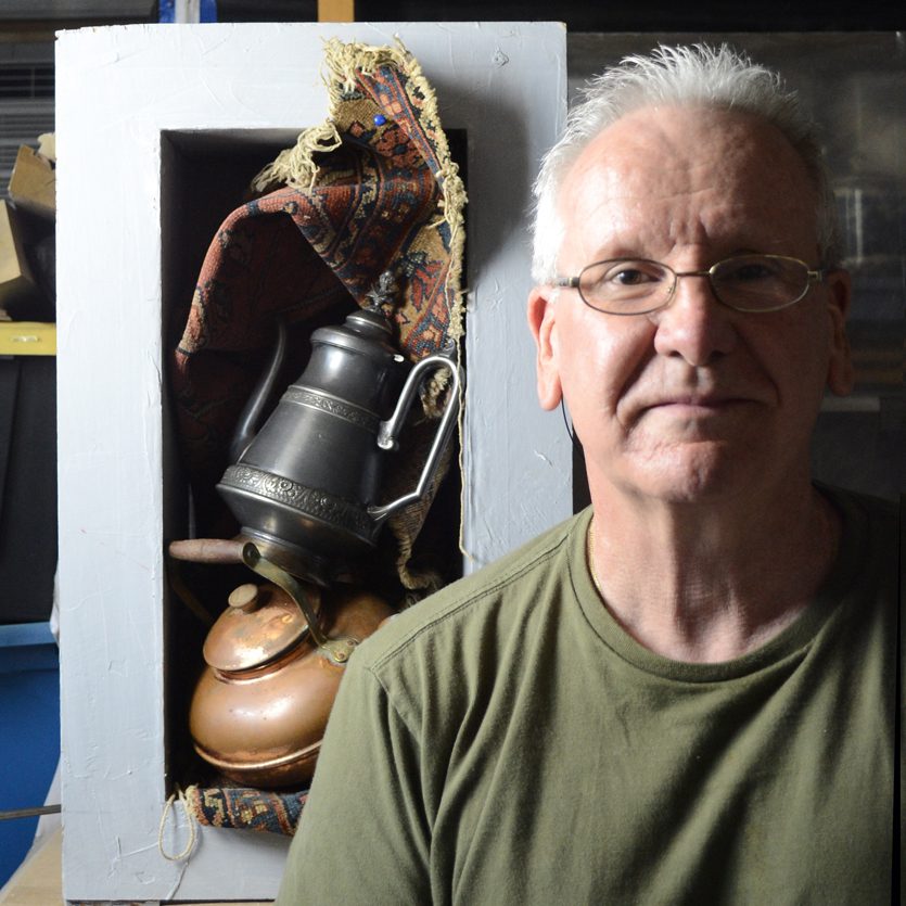 A man in glasses stands beside a vintage coffee pot and kitchenware.