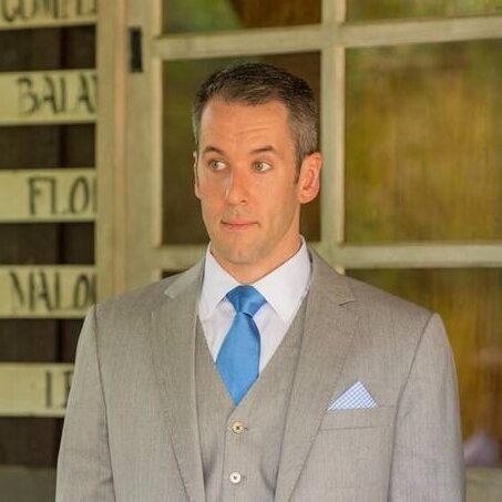 Man in a gray suit and blue tie standing indoors.